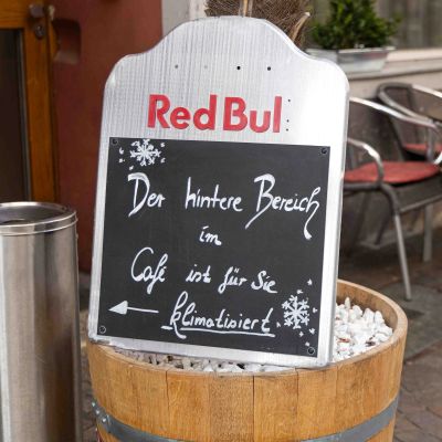 Aufsteller vor einem Café mit der Kreideaufschrift: „Der hintere Bereich im Café ist für Sie klimatisiert“ mit Pfeil nach links und Schneeflocken-Zeichnungen.
