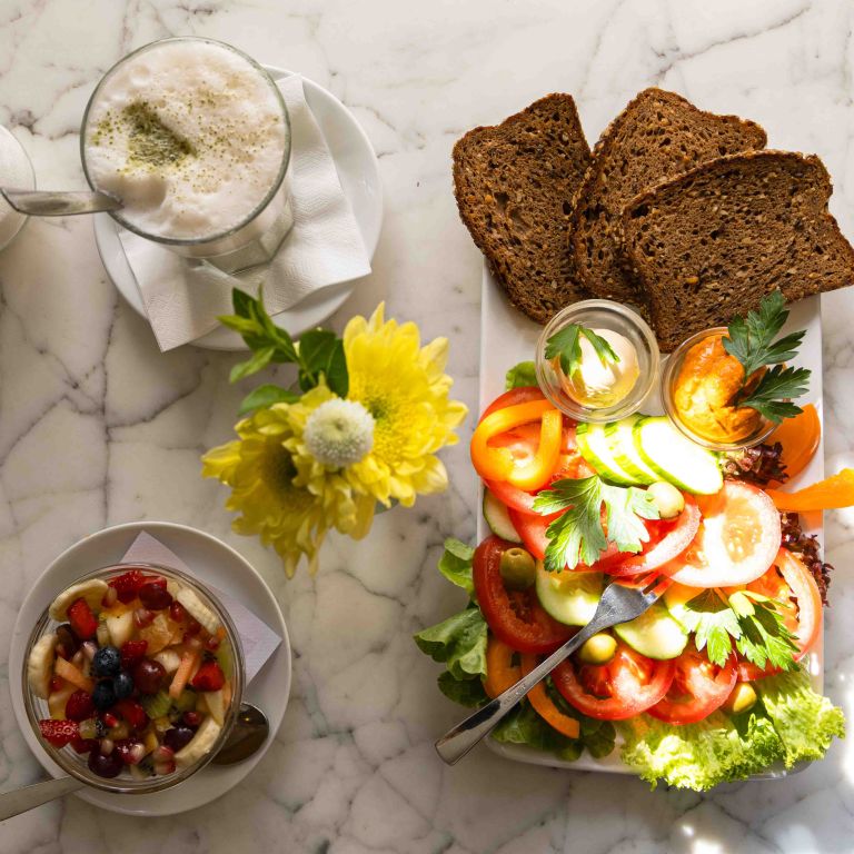 Draufsicht auf einen Salat, Dips, drei Scheiben Vollkornbrot, Obstsalat, Matcha Latte und einer gelben Blüte.